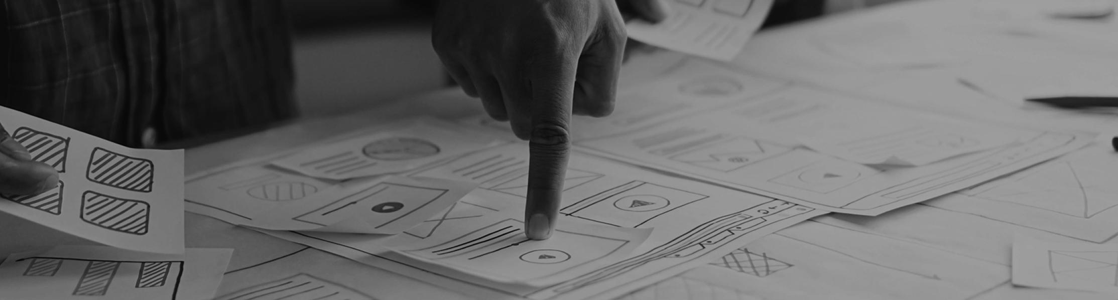 Graphic showing papers with designs on a desk and a hand pointing at a design 