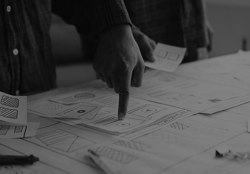 Graphic showing papers with designs on a desk and a hand pointing at a design 